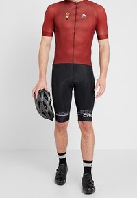 Rød kortærmet cykeltrøje med lynlås foran, sorte polstrede shorts, sorte cykelsko og en hjelm i hånden. Detaljer inkluderer logo og mønstre.