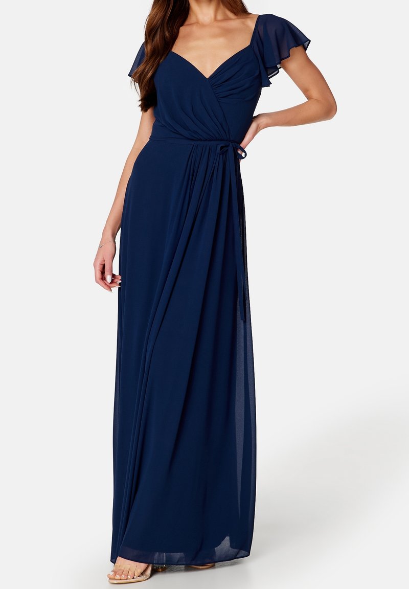 Femme portant une robe portefeuille bleu marine à manches flottantes et décolleté en V, posant avec une main sur la hanche.