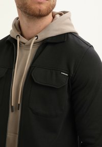 Uomo che indossa un cappuccio beige sotto una giacca nera con zip, dotata di tasche a pattina sul petto e etichetta "GABBIANO" sulla tasca sinistra.