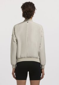 Jachetă bomber de culoare bej deschis, cu o textură netedă, manșete și tiv zimțat, și un orificiu mic de ventilație la spate. Pur tată pe deasupra unor pantaloni scurți negri.