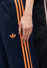 Χέρι που ακουμπάει πάνω σε μπλε-ναυτικό παντελόνι Adidas με πορτοκαλί ρίγες και λογότυπο, δίπλα σε άλλο άτομο που φοράει μαύρο παντελόνι.