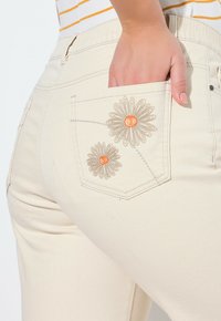 Pantalones de mezclilla beige con bolsillo trasero que presenta bordados florales en gris y naranja. Textura suave y diseño ajustado con una cintura de altura media.