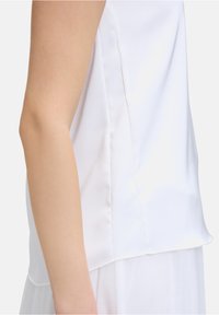 Weißes ärmelloses Shirt aus glattem Stoff mit leicht abgerundetem Saum und seitlichen Nähten. Der Stoff ist leicht und hat eine weiche Textur.