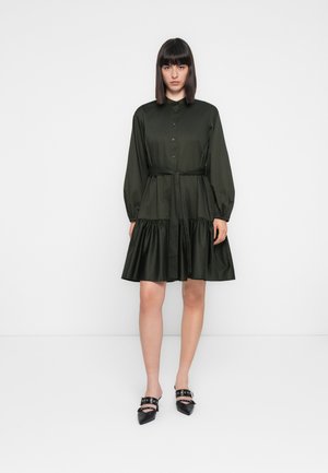 Vrouw in een knielang zwart jurkje met lange mouwen, knoopsluiting aan de voorkant, een strik rond de taille en zwarte, puntige schoenen met banden.