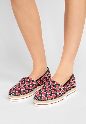 Stopy w butach na platformie typu slip-on z czarno-czerwonymi pikselowymi wzorami serc oraz białymi podeszwami wykończonymi jutą na szarym tle.