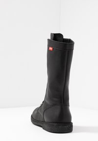 Black leather knee-high boot with a textured finish, featuring a narrow shape, sturdy sole with a slight tread, and a small red label.