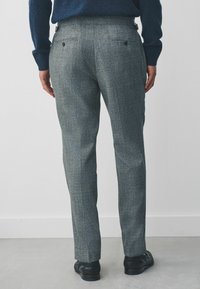 Graue, strukturierte Hose mit schmaler Passform, ausgestattet mit zwei Gesäßtaschen und subtilen Nahtdetails. Kombiniert mit schwarzem Schuhwerk.