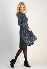 Marinblå klänning med prickmönster, figurnära midja, långa ärmar och utsvängd kjol. Modellen bär svarta skor och strumpbyxor.