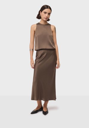 Femme aux cheveux courts et foncés portant un haut marron en satin sans manches et une jupe assortie longueur cheville avec des chaussures noires pointues.