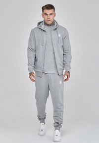 Sudadera gris con capucha, cremallera frontal y cordones, combinada con pantalones deportivos a juego. Ambos tienen un logo discreto; la tela parece suave y texturizada.
