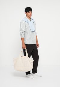 Bolsa de lona beige con dos asas resistentes, forma rectangular y textura suave. El modelo lleva una camisa a rayas y pantalones azul marino.