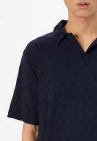 Μπλέ ναυτικού πόλο πουκάμισο φτιαγμένο από υφασμάτινη υφή με καρό μοτίβο, διαθέτοντας γιακά και κοντά μανίκια.