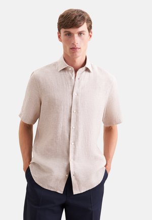 Ung mand iført en beige kortærmet skjorte med knapper og mørkeblå bukser, stående med hænderne i lommerne mod en hvid baggrund.