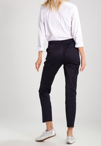 Námořnické kalhoty na míru se slim fit, hladkou texturou a kapsami, v kombinaci s bílou košilí na knoflíky a stříbrnými teniskami.