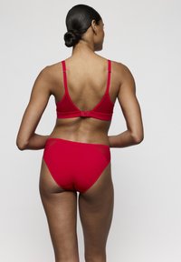 Vrouw met donker haar in een knot, gekleed in een bijpassende rode beha en hoge taille slip, staand met haar rug naar de camera op een effen achtergrond.