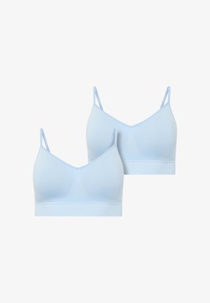 Smiling woman with long wavy hair wearing a light blue sports bra and black underwear, standing barefoot on a plain background.