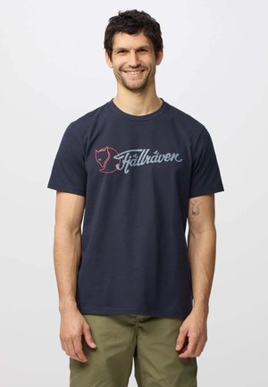 Homme souriant portant un T-shirt bleu marine avec un logo de renard rouge et le mot "Fjällräven" en écriture bleu clair, et un pantalon vert olive.