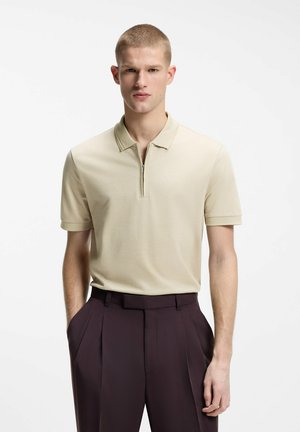 Mężczyzna w beżowej polówce z krótkim rękawem i suwakiem oraz ciemnych plisowanych spodniach z wysokim stanem, stoi z rękami w kieszeniach.