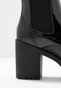 Bottines en cuir verni noir avec un talon épais, présentant des panneaux latéraux élastiques et des détails de couture décoratifs. Fini texturé sur le talon.