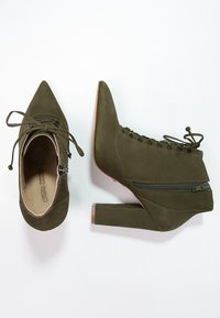 Botas de tobillo verde oliva con punta afilada y superficie texturizada. Cuentan con una cremallera lateral y detalles de encaje en la parte superior. Tacón cuadrado.