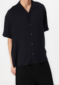 Camisa negra de manga corta hecha de tejido ligero, con cuello de campamento, frente abotonado y un corte holgado. Textura suave, sin patrones.