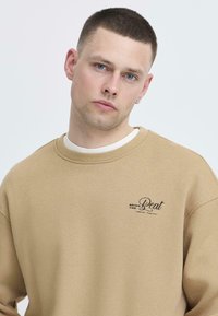 Giovane uomo con capelli corti e scuri e occhi azzurri, che indossa una felpa beige con la scritta "Bring the Beat" sul petto, su uno sfondo bianco uniforme.