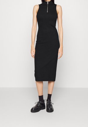 Ärmelloses schwarzes Midikleid mit hohem Kragen und Reißverschluss vorne, kombiniert mit schwarzen Socken und massiven schwarzen Schnürschuhen.