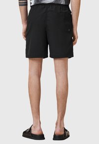 Costume da bagno neri con vita elasticizzata, una tasca sul retro e una texture morbida. Indossati con sandali neri, che mostrano gambe abbronzate.