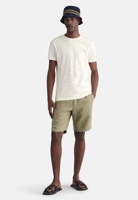 Hvid t-shirt med korte ærmer, olivengrønne shorts og sorte sandaler. Model iført en marineblå bucket hat med en stripes accent.