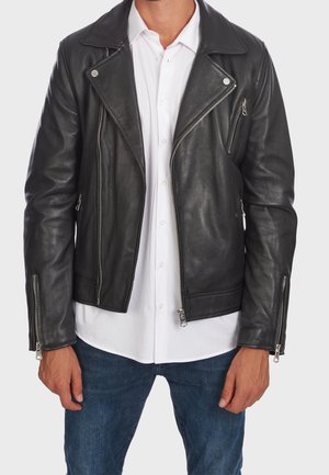 Homme portant une veste de motard en cuir noir par-dessus une chemise blanche boutonnée et un jean bleu, debout devant un fond uni.
