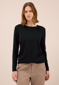 Kvinna med axellångt ljusbrunt hår bär en svart långärmad skjorta och beige joggerbyxor, stående mot en ljus bakgrund.