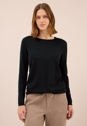 Femme avec des cheveux châtains clair jusqu'aux épaules, portant un t-shirt noir à manches longues et un pantalon beige à cordon de serrage, se tenant devant un fond clair.