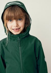 Grön vattentät jacka med dragkedja framtill, fast huva och vita detaljer. Har en cirkulär logotyp på ärmen och en slät yta.