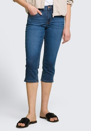 Frau trägt blaue Jeans mit mittlerer Taille und verkürztem Bein, dazu eine beige Jacke, weißes Oberteil und schwarze Sandalen mit offenem Zehenbereich, steht vor einfarbigem Hintergrund.