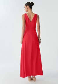 Punane varrukateta maksikleita V-kaelusega tagaküljel, sileda tekstuuriga ja lukuga tagant. Kandnud alasti toonides kõrgete kontsadega kingadega.