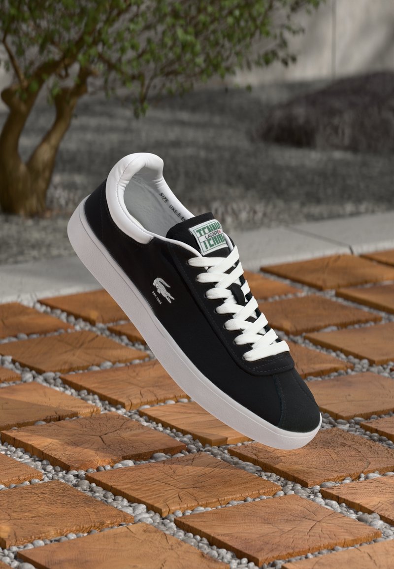 Baskets Lacoste noir et blanc avec lacets blancs et logo, présentées sur des carreaux carrés en bois avec de petites pierres grises en extérieur.