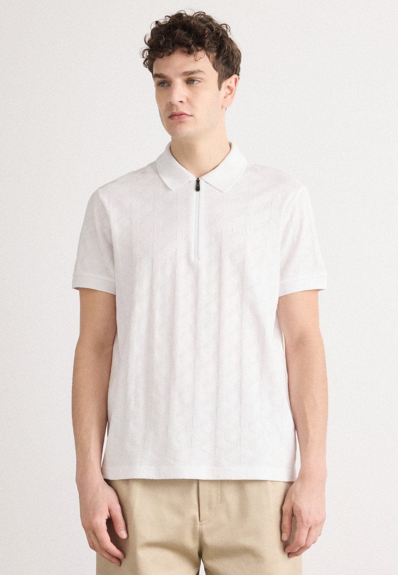 Camisa polo blanca con un patrón geométrico texturizado, mangas cortas y cierre de cremallera en el cuello. Presenta un logotipo sutil en el pecho.