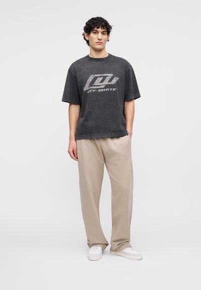 Νεαρός άνδρας με σγουρά μαλλιά στέκεται φορώντας μια ξεθωριασμένη μαύρη μπλούζα με σχέδιο, μπεζ παντελόνι και λευκά αθλητικά παπούτσια, πάνω σε απλό φόντο.