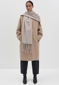 Cappotto di lana beige dal taglio morbido, maniche lunghe, tasche laterali. Sciarpa grigia con frange drappata attorno al collo. Stivaletti neri alla caviglia.
