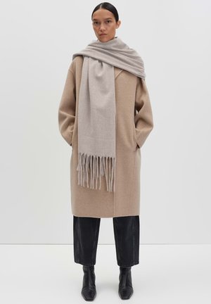 Beige wollen jas met een relaxte pasvorm, lange mouwen, zijzakken. Grijze sjaal met franjes draped om de hals. Zwarte enkellaarsjes.