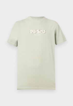 Hellbeiges T-Shirt mit kurzen Ärmeln und der Aufschrift "GUESS EST. 1981" in Weiß auf der Brust, präsentiert vor einem einfarbigen Hintergrund.