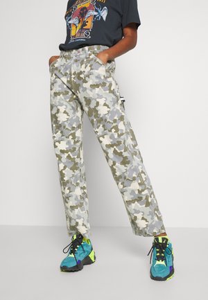 Person wearing green and gray camouflage pants, graphic black t-shirt, and blue and neon yellow sneakers standing with hands in pockets.