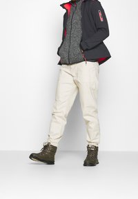 Personne portant un pantalon de jogging beige, des bottes à lacets vert olive, un sweat à fermeture éclair gris et une veste noire avec un intérieur et des accents rouges.