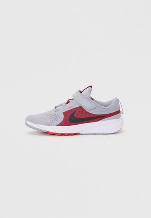 Chaussure de sport avec une tige en mesh gris agrémentée d'accents rouges, un swoosh noir de Nike, une sangle Velcro et une semelle blanche texturée avec des détails rouges.