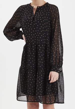 Svart klänning med prickar, långa ärmar, genomskinligt tyg, knäppt placket, vågig kjol och rund halsringning; har ljusbeige prickar.