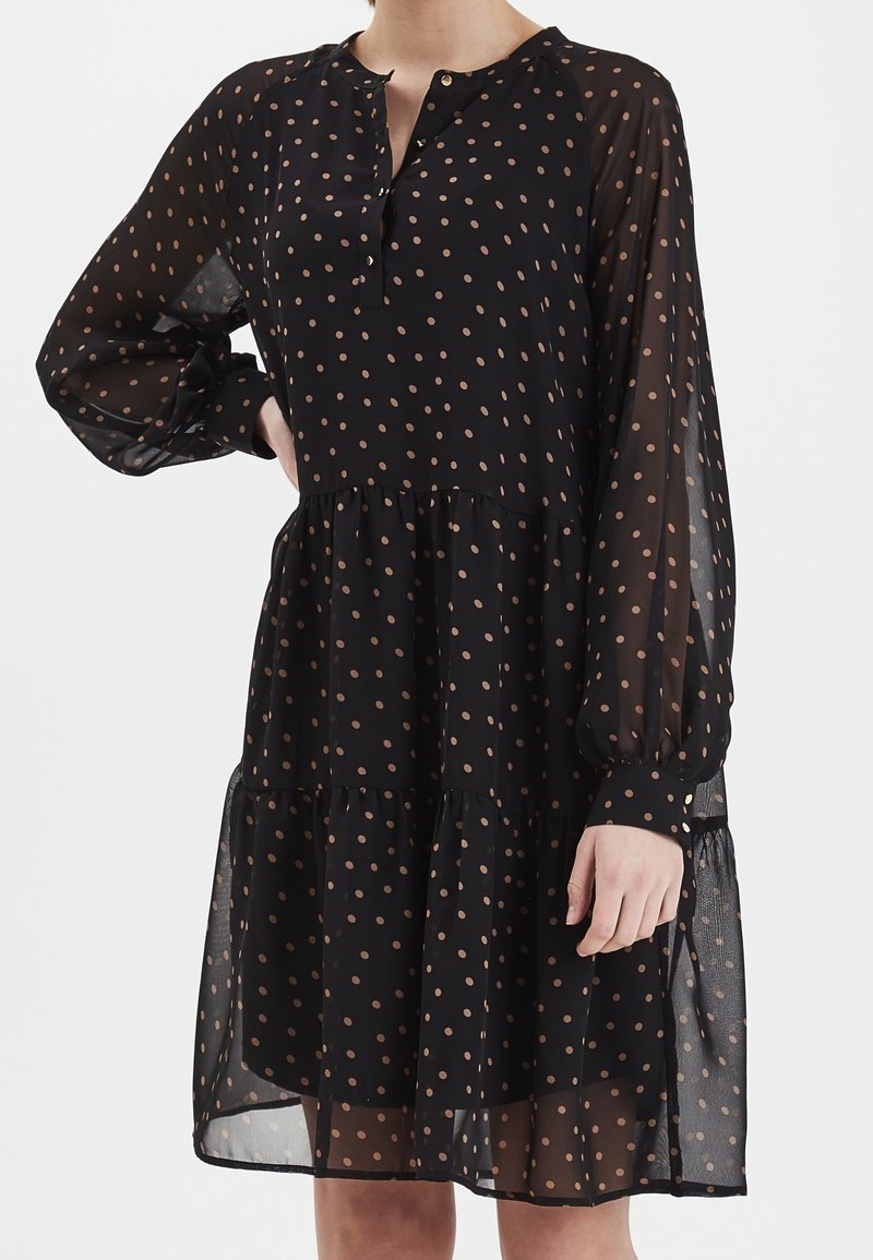 Černé šaty s puntíky, dlouhými rukávy, průsvitnou tkaninou, klopovým zapínáním, vrstevnatou sukní a kulatým výstřihem; mají světle béžové puntíky.