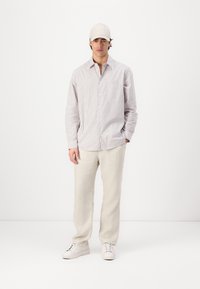 Chemise à rayures dans des couleurs claires, associée à un pantalon en lin beige et des baskets blanches. Une casquette de couleur claire complète la tenue.