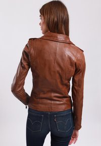 Braune Lederjacke mit einer taillierten Passform, breitem Kragen und Reißverschlussärmeln. Getragen über dunkelblauen Jeans mit Taschenverzierungen.