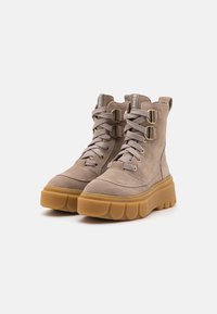 Botas de tobillo de ante marrón claro con suela de goma gruesa, con cordones en la parte delantera y dos hebillas metálicas en los lados.