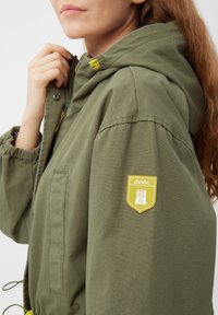 Giacca a vento verde oliva con cappuccio realizzata in un tessuto leggero, dotata di cappuccio regolabile, polsini elastici e un'etichetta gialla sulla manica.